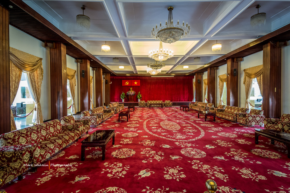Large ornate room with red patterned carpet, gold and red upholstered sofas along the sides, chandeliers hanging from the ceiling, and red curtains with a podium beneath communist and Vietnamese flags at the far end.