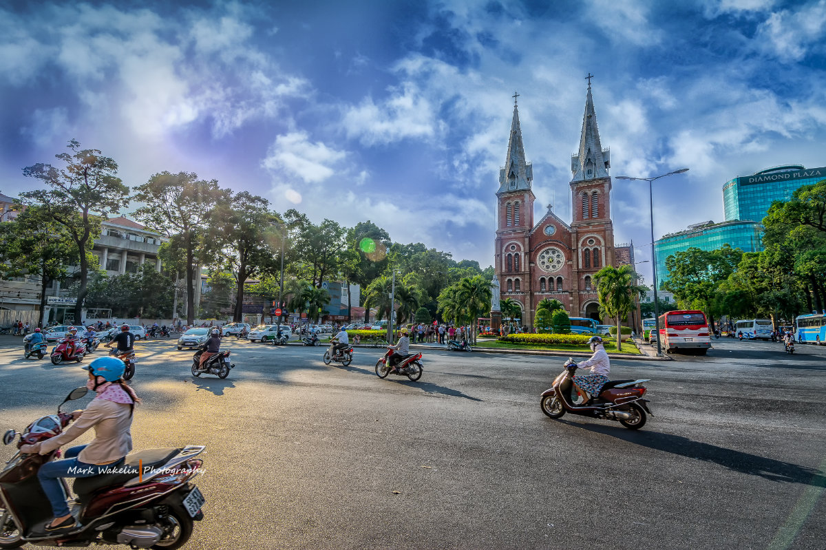 Busy street scene in front of the Notre-Dame Cathedral Basilica of Saigon with people on scooters and a bright sky.