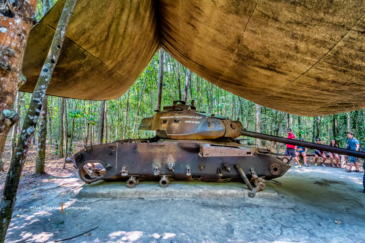 Rusty old tank displayed under a large canopy in a dense forest with people sitting and walking nearby.