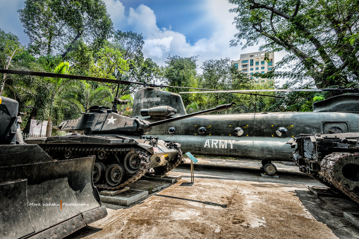 U.S. Army military helicopter and tank displayed outdoors surrounded by trees and a building in the background.