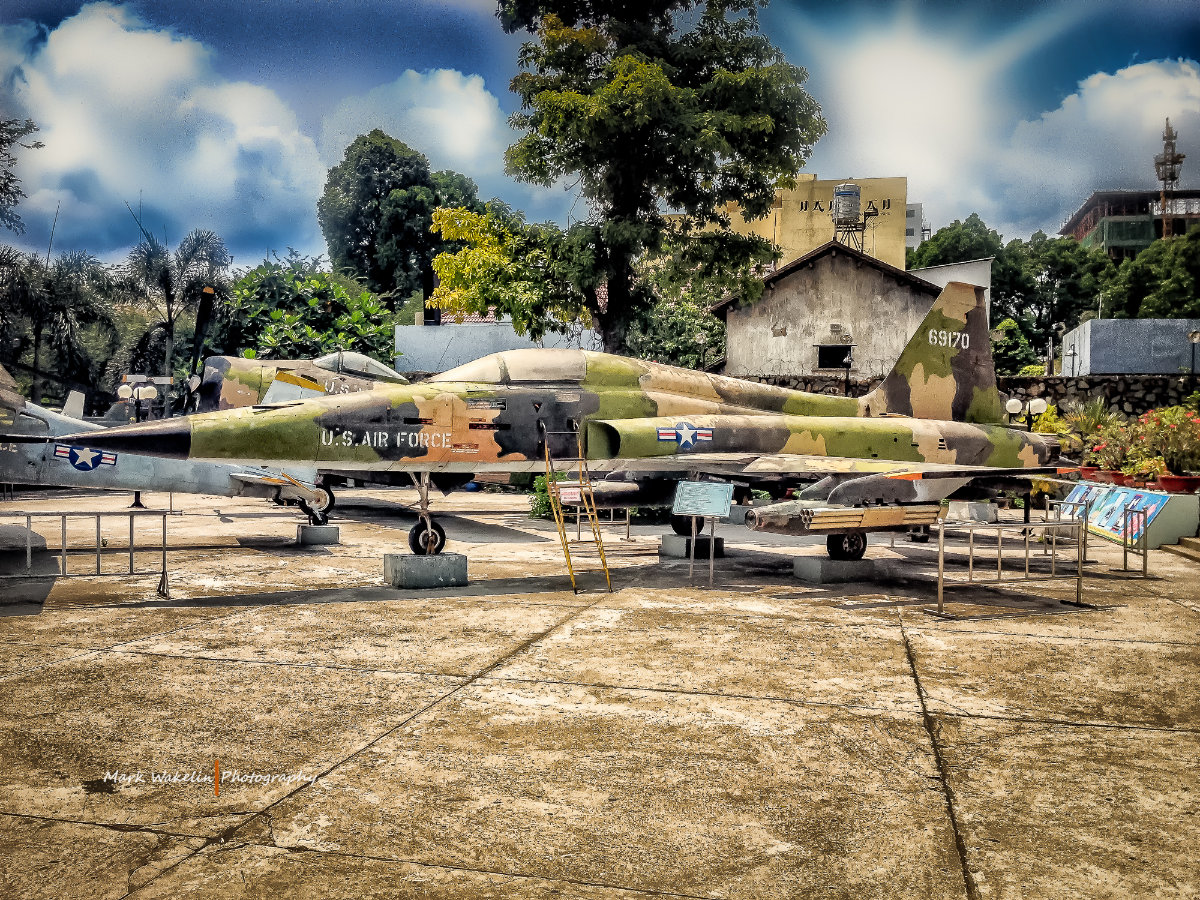 Camouflaged U.S. Air Force fighter jet displayed outdoors with a yellow ladder resting against it and trees and buildings in the background.