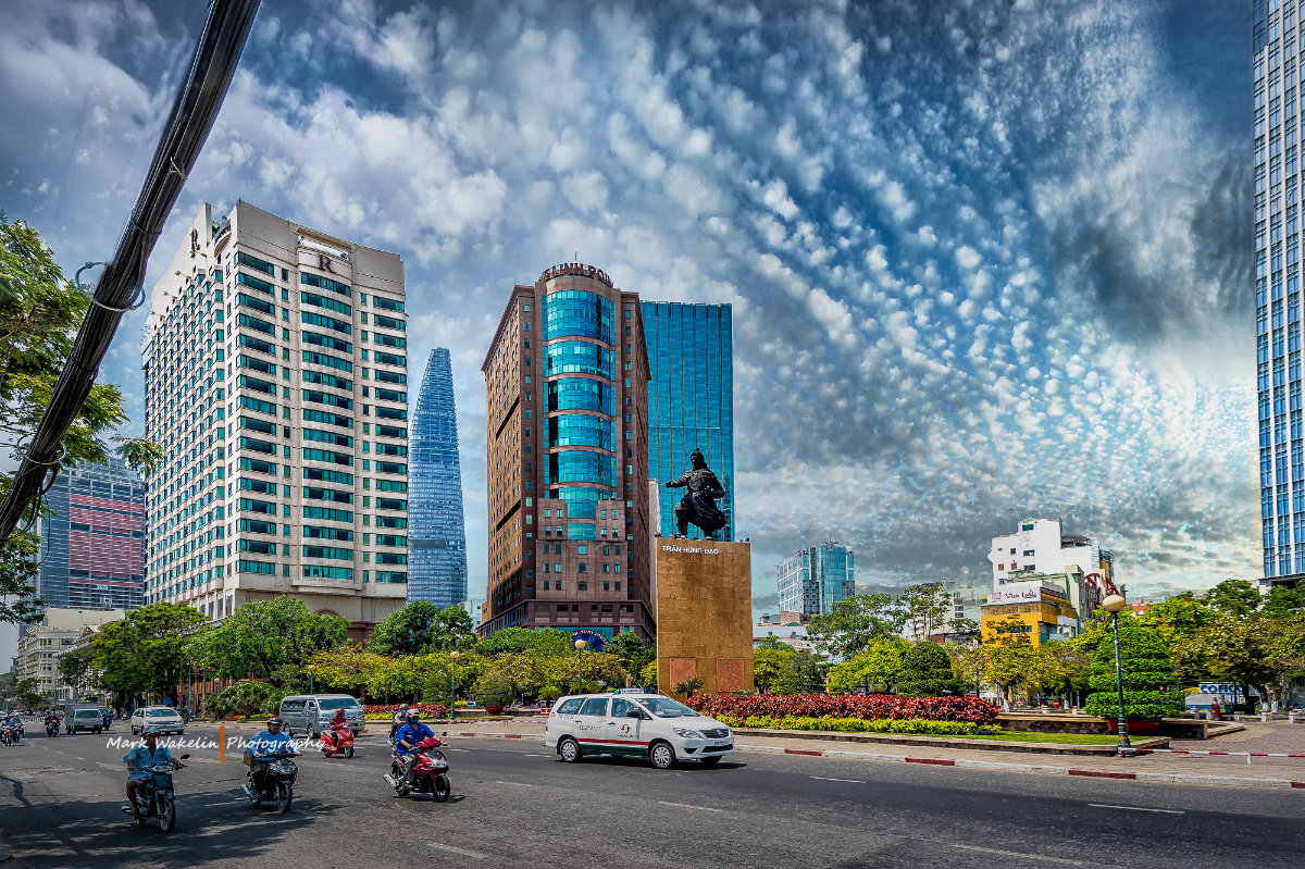 Urban street with motorcycles and cars in front of modern buildings and a statue under a dramatic cloudy sky.
