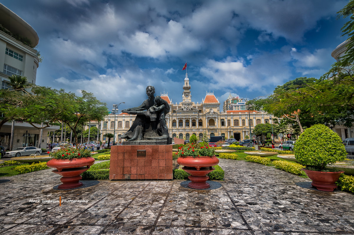 Statue of a seated man holding a child in a landscaped garden square with historic colonial-style building and Vietnamese flag in the background under a partly cloudy sky.