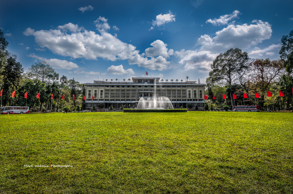 Wide view of a large green lawn leading to a rectangular building with a water fountain in front, flanked by red flags and trees under a partly cloudy blue sky.
