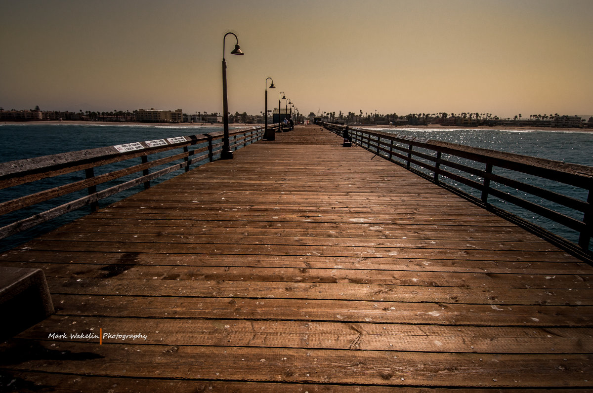 Wide wooden pier with lampposts extending over blue sea towards a distant shoreline with buildings and palm trees under a hazy sky.