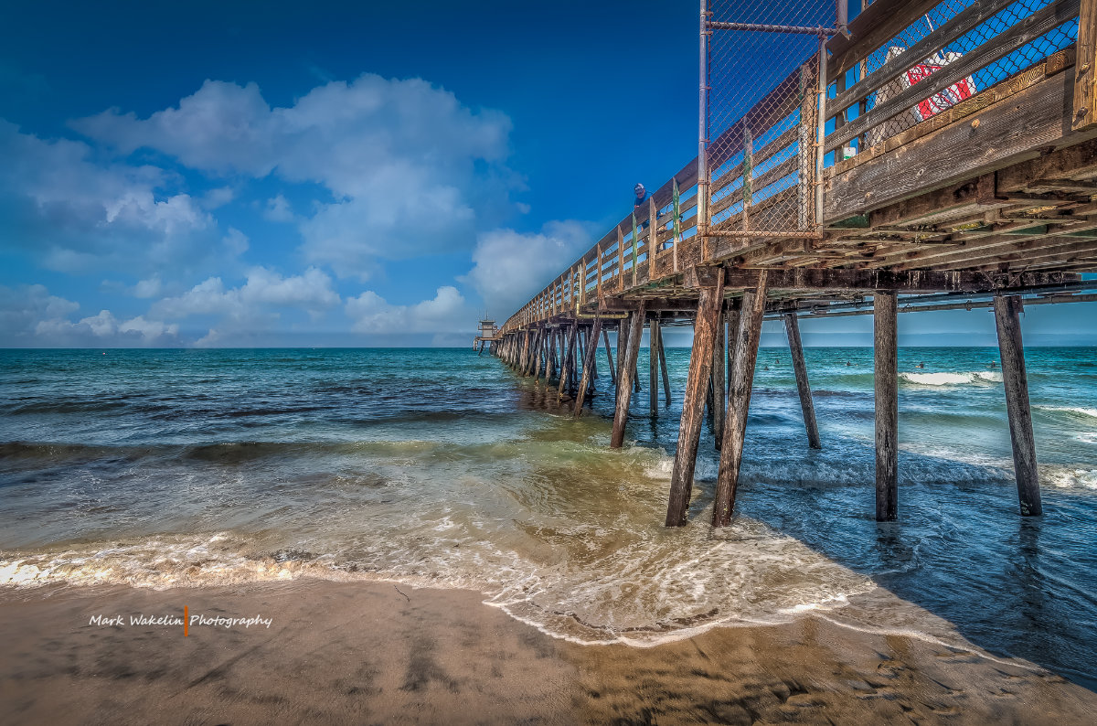 Wooden pier extending over the ocean with waves gently breaking on the sandy beach under a partly cloudy blue sky.