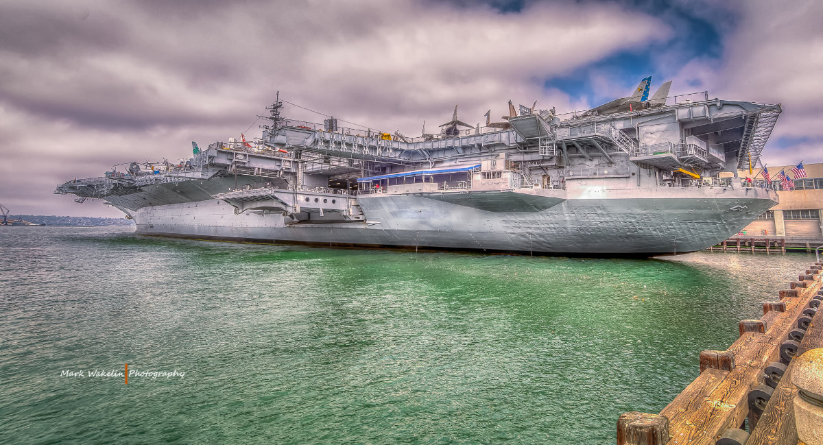 Large grey aircraft carrier docked at a pier with a cloudy sky overhead and calm green water surrounding it.