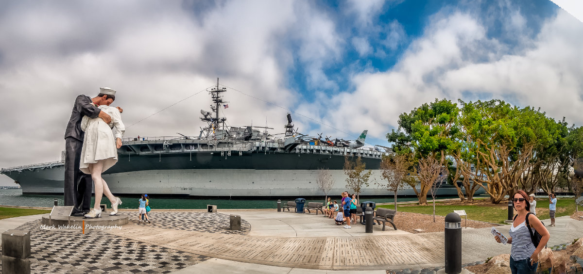 Visitors at a waterfront park with the iconic 'Unconditional Surrender' statue and a large aircraft carrier in the background under a partly cloudy sky.