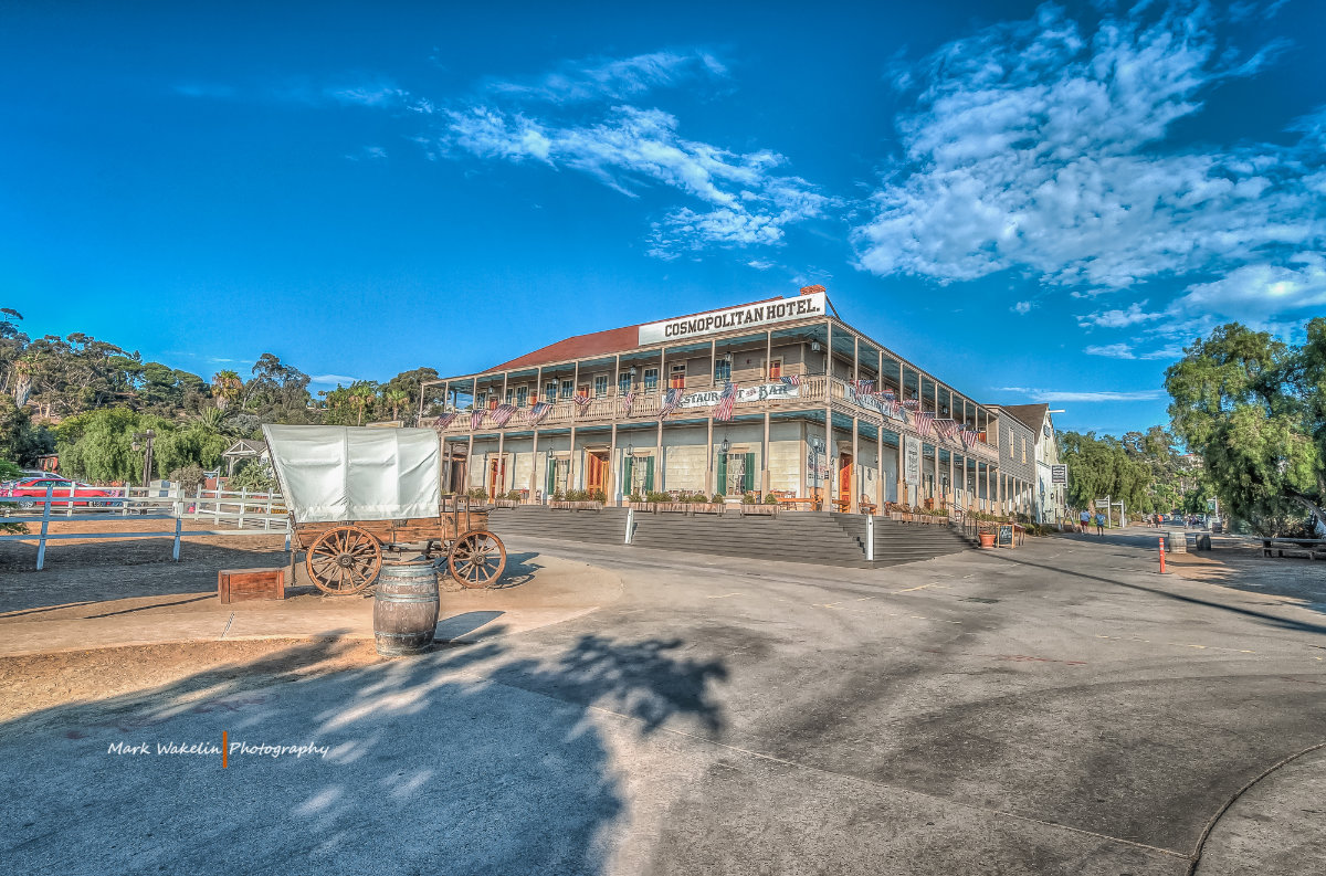 Historic Cosmopolitan Hotel with covered porch, American flags, and a wooden covered wagon and barrel in front under a blue sky.