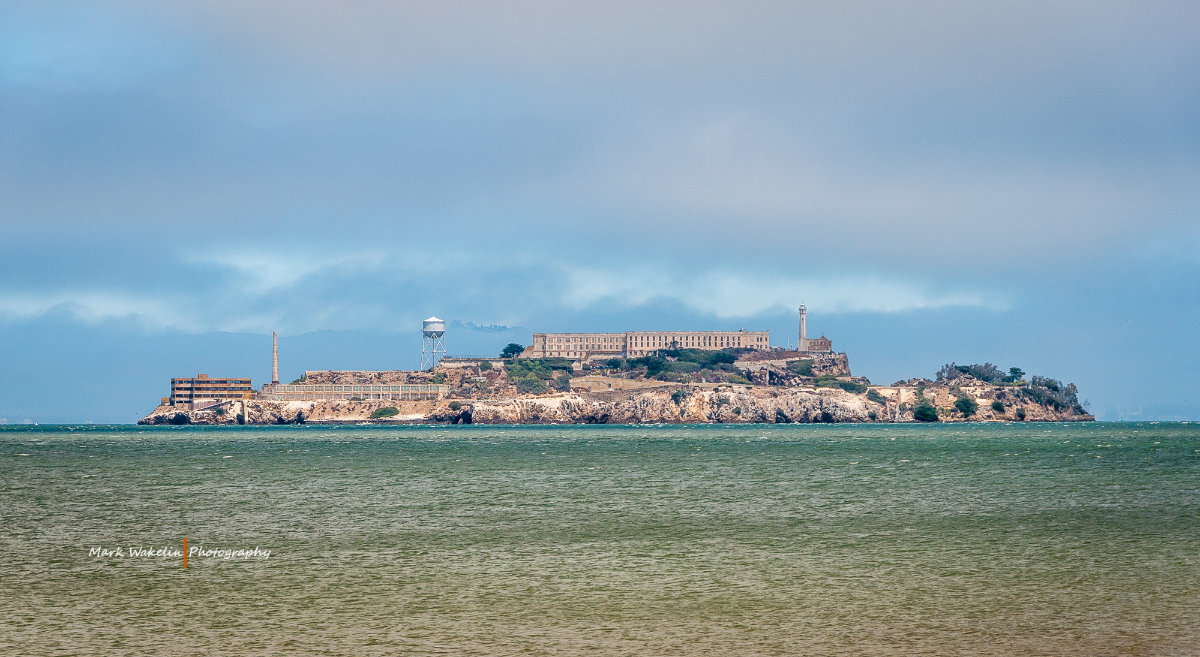 Distant view of Alcatraz Island with historic prison buildings and lighthouse surrounded by ocean under a cloudy sky.