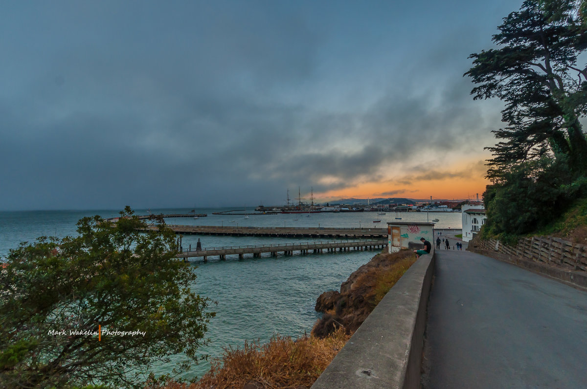 Sunset view of a waterfront with piers extending into the water, a few people walking along a path, and a large tree on the right side.