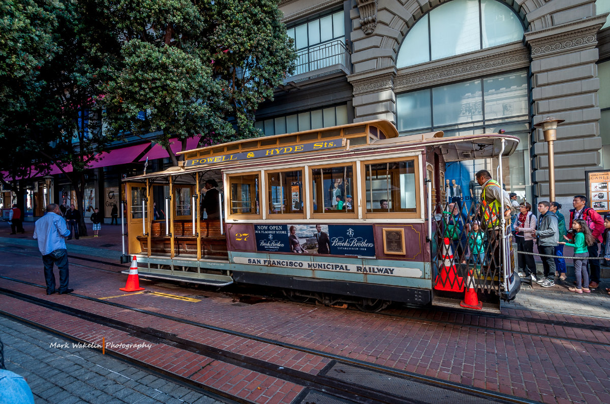 San Francisco cable car on Powell & Hyde Sts. with passengers boarding and waiting beside it on a city street.