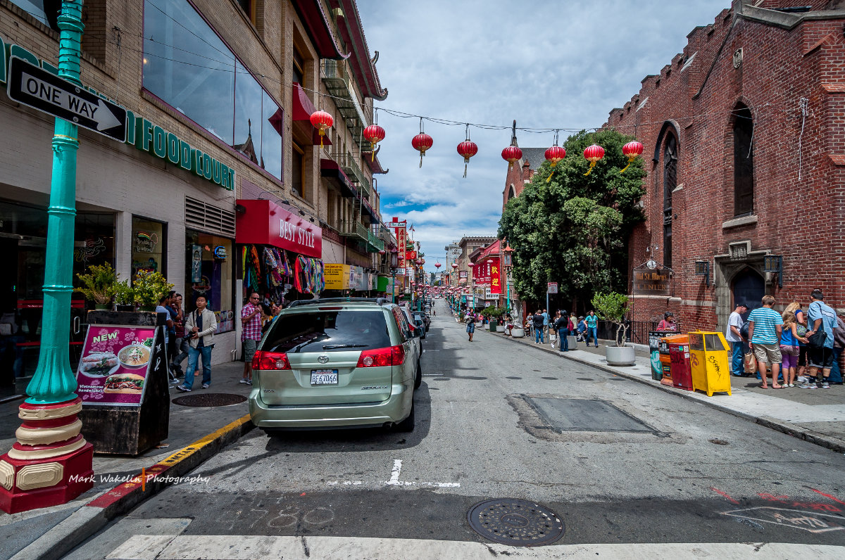 Street view of Chinatown in San Francisco with parked cars, pedestrians, red lanterns hanging above, and various shops including a food court and souvenir stores.