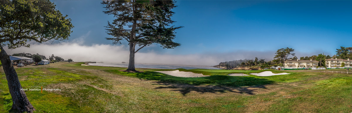 Wide panoramic view of a golf course with green fairways, sand bunkers, tall trees, buildings on the right, and a fog bank receding in the background under a clear blue sky.